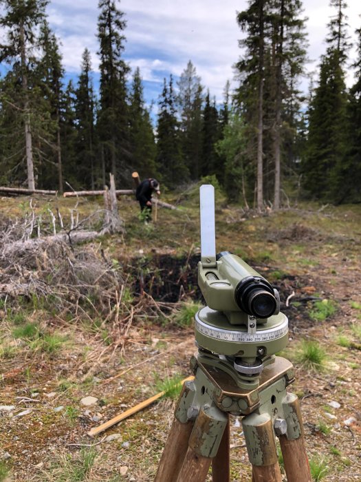 Nivåinstrument i förgrunden med person som mäter ut marken för byggprojekt i skogsmiljö i bakgrunden.