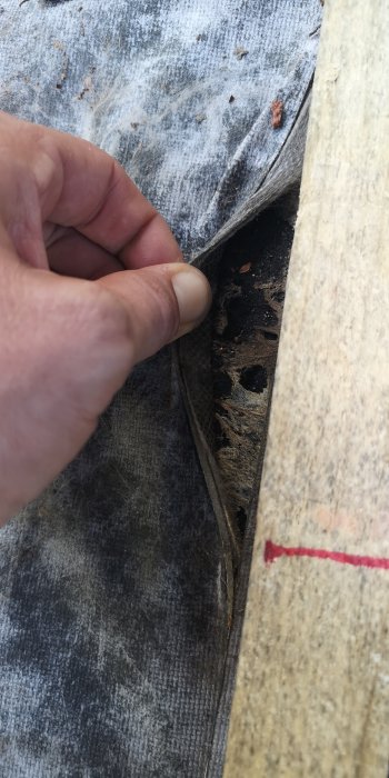 Hand lyfter på en underlagsduk på ett tak som visar en dåligt utförd skarv med minimal överlappning och underliggande trästruktur.