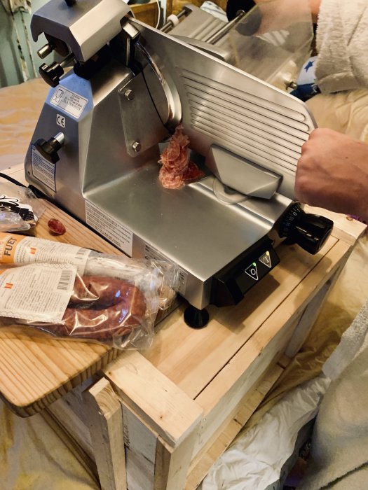 Person som använder en elektrisk skärmaskin för att skiva salami på ett träbord.