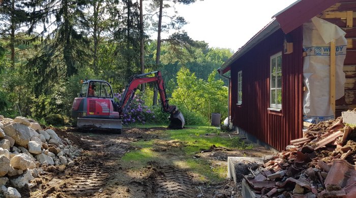 Grävmaskin som gräver i trädgård nära hus för anläggning av markbädd, med hög med sten och grushög i förgrunden.