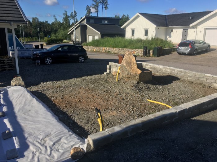 Byggarbetsplats med öppen glugg i mur för lastbil, grushög och stenblock, i ett bostadsområde.