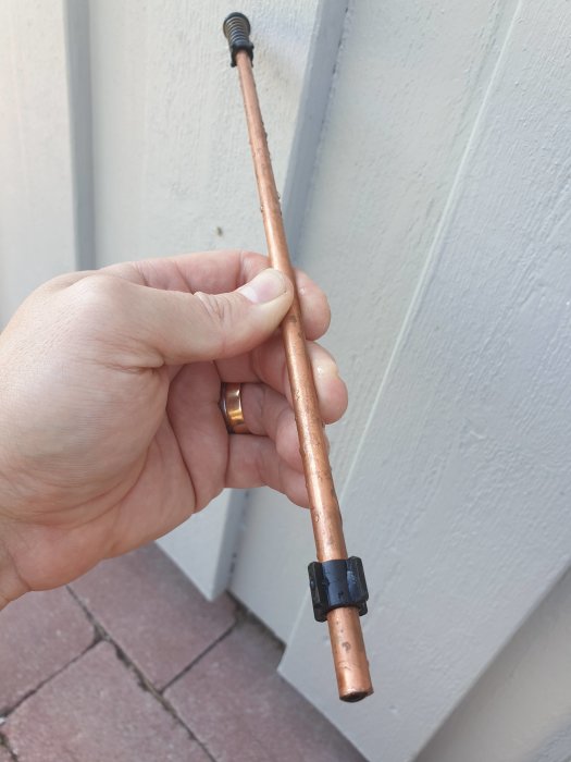 Hand håller en koppar vattenutkastare med svart justerbar ring nära kameran mot en husvägg.