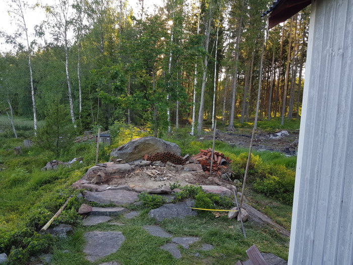 Tom plats i trädgård med stenar och ved, förberedd för gjutning av platta, omgiven av grönska och skog.