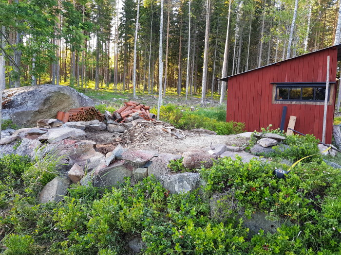 Röd vedbod bredvid en hög med stenar och en stor sten, gammalt byggmaterial på marken i skogsmiljö.