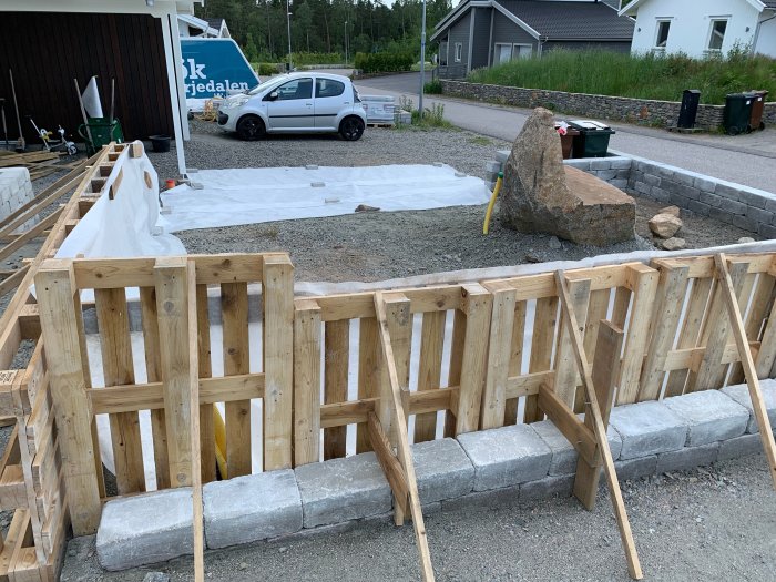 En träramkonstruktion byggd runt en låg stenmur för förberedelse av större jordleverans med markduk framlagd på marken.