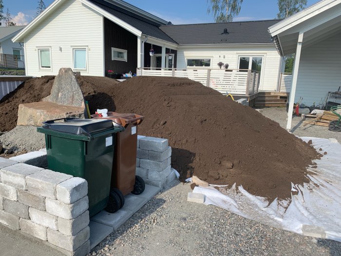 Nylevererad hög av matjord framför ett hus, med sophantering och byggmaterial i förgrunden.