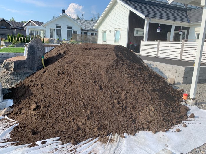 En stor hög med matjord placerad noggrant på en trång yta vid ett hus med murar och carport i bakgrunden.