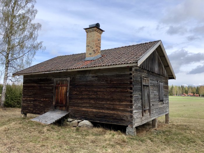 Gammal fäbod med trästomme och tegeltak omgiven av öppet landskap och några träd.