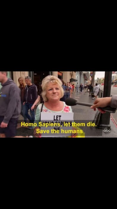 Kvinna håller skylt med texten "LET Homo Sapiens die. Save the humans" medan hon blir intervjuad på gatan.