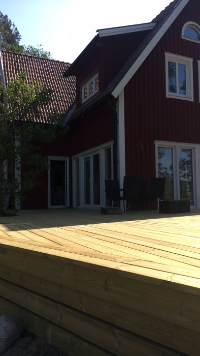 Nybyggd träaltan framför ett rödmålat hus med vita fönster och terrassdörrar, möblerad med svarta utestolar.