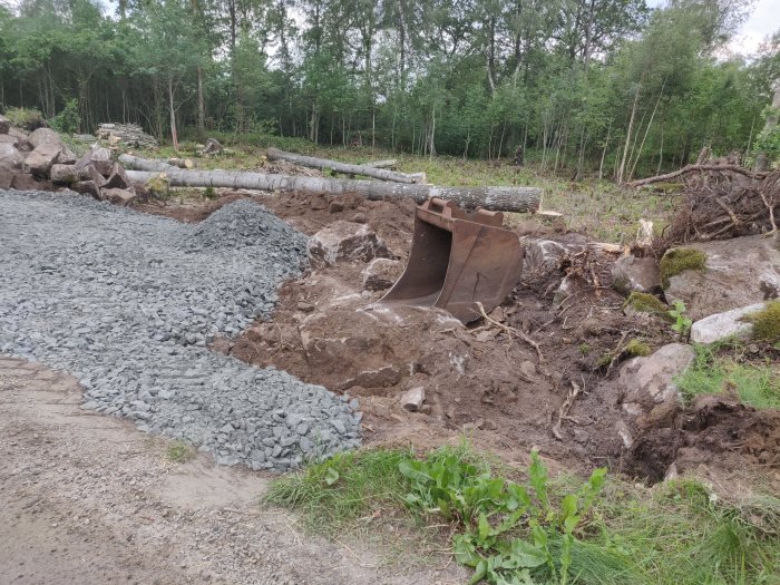 Nygrusad parkeringsplats vid byggarbetsplats med skog i bakgrunden och en grävmaskinskopa i förgrunden.