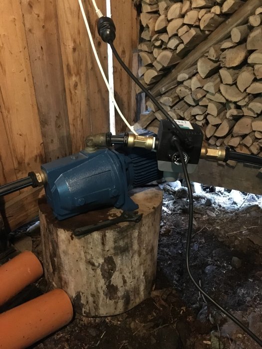 Vattenpump placerad på stubbe i vedförråd, ansluten med slangar och kablar, för trädgårdsbevattning.