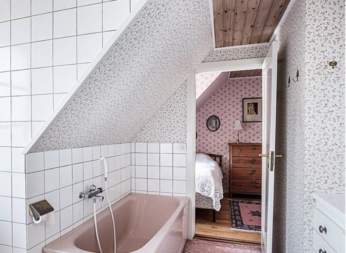 Rosa badkar i ett kaklat badrum med öppen dörr som leder till ett sovrum med blommig tapet och träbyrå.