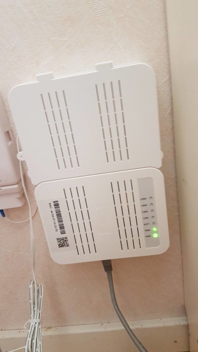 Äldre vit router monterad på vägg med synliga kablar och gröna lysdioder indikerar fungerande anslutning.