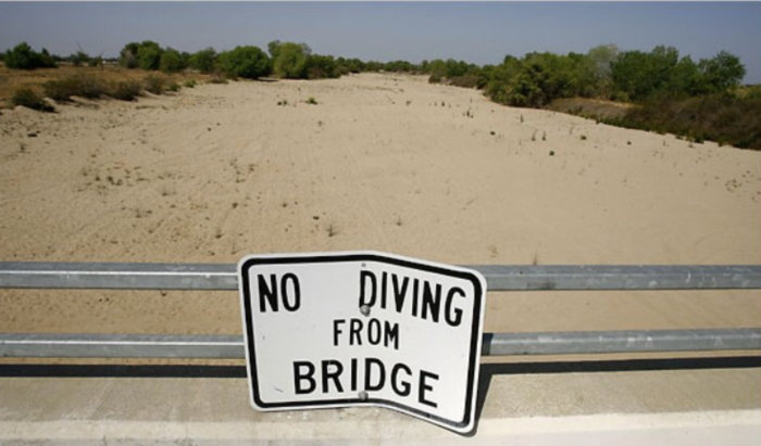 Ironisk "No Diving from Bridge"-skylt vid torrlagd flodbädd som belyser klimatförändringars effekt.