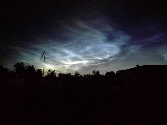 Mörk himmel med skimrande blått och vitt ljus över silhuetter av byggnader och träd under midsommarnatten.