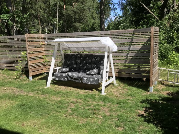 En hammock med mönstrade kuddar täcks av ett vitt takdukskydd för att skydda mot regn i en grön trädgård.