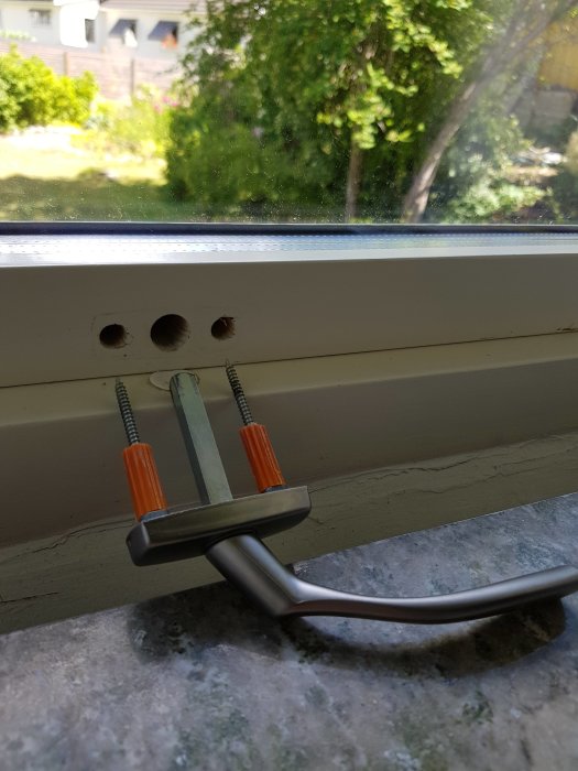 Ett löst vridfönsterhandtag med synliga skruvar på fönsterkarmen, med en suddig trädgård i bakgrunden.