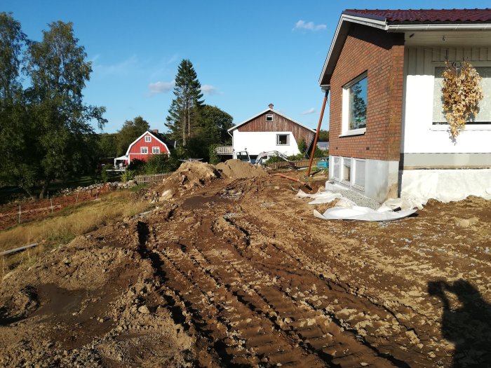 Hus med pågående dräneringsarbete, stora jordhögar och spår efter grävmaskin i förgrunden.