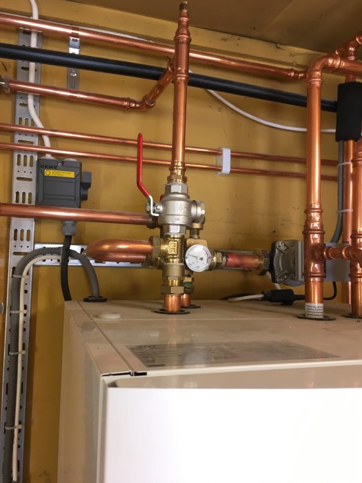 Rörinstallation och mätare ovanför en vit apparat, med kablar och väggfästen i bakgrunden.