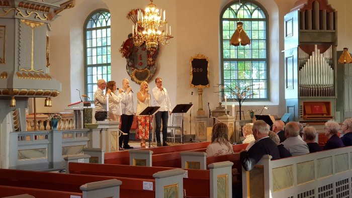 Fyra personer sjunger framför mikrofoner inne i en kyrka medan en publik lyssnar.