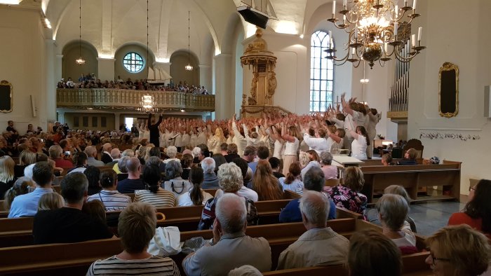 Kör framför publik i kyrka med händerna uppsträckta och orgel i bakgrunden.