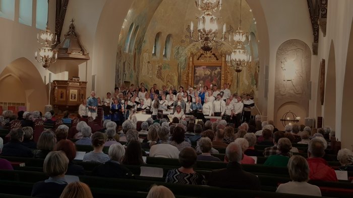 Kör framför publik i kyrksal med målade väggar och kristallkronor.