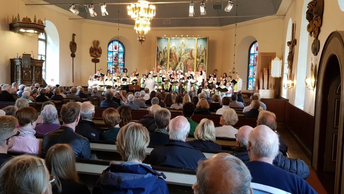 Kör framför publik i kyrka med dirigent och målning i bakgrunden.