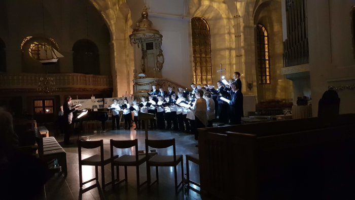 Kör upplyst av notlampor framför sig under en konsert i en mörklagd kyrka.