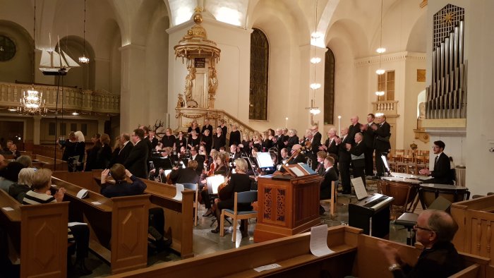 Kör och orkester uppträder i kyrka med publik och kyrkorgel i bakgrunden.