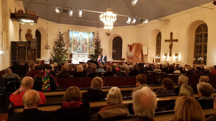 Interiör av en kyrka under en gudstjänst med besökare som sitter och lyssnar på prästen.