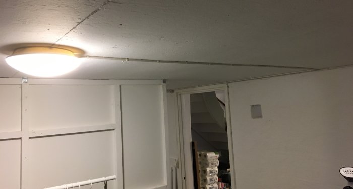 Plafond med tänd ljus i källartvättstuga med vita väggar och synlig skiljevägg.