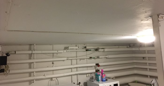En tvättstuga med vita rör längs taket och en tänd plafond som ger otillräckligt ljus.