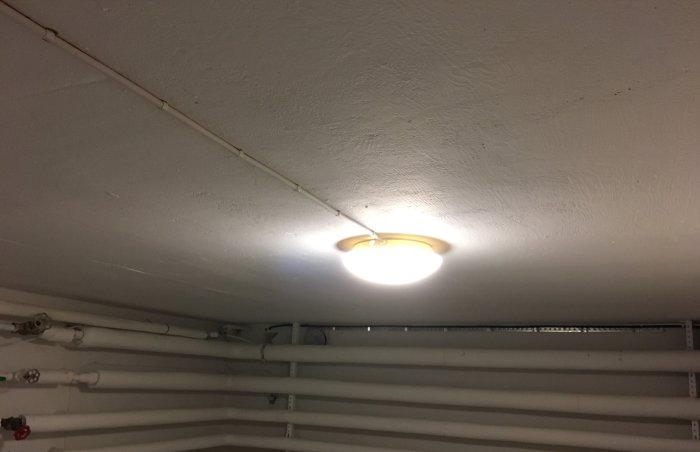 Källarbelysning med tänd plafond på en vitmålad takyta, omgiven av vita rör och kablar.