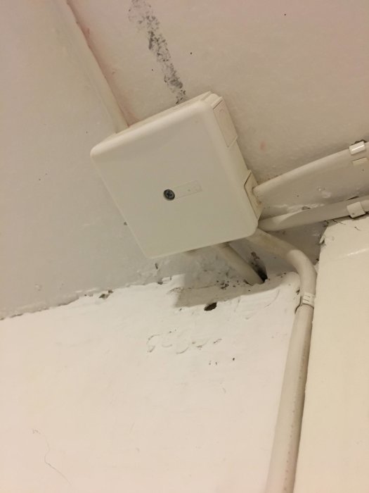 En vit elektrisk kopplingsdosa med rörinstallationer i ett hörn av ett rum med vita väggar.