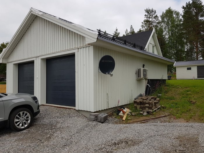 Vit garagebyggnad med svarta garageportar, grusuppfart och en parabolantenn på väggen.