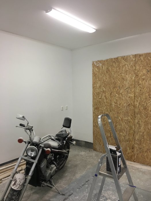 Renoverat rum med nyligen installerad belysning, ommålad vägg och plywoodtäckt dörröppning, motorcykel och stege inuti.