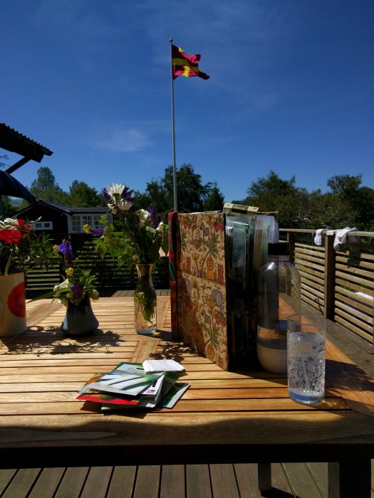 Ett soligt trädäck med fröpåsar, en vattenkaraff, ett glas vatten och en vas med blommor, i bakgrunden en svensk flagga vajar.