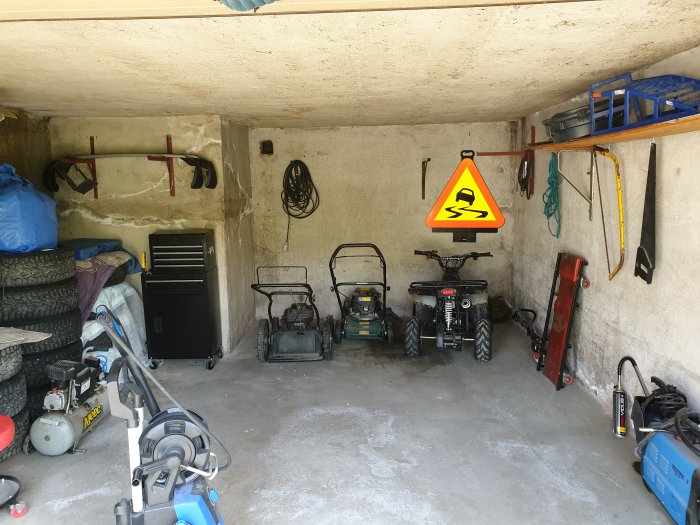 Bild av ett garage med ojämna väggar och tak, verktyg hängande på väggen och olika trädgårdsredskap och däck lagrade.