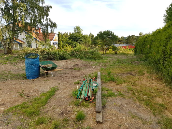 Röjt hörn av trädgård med skottkärra, säck och grenar på marken, i förortsområde.