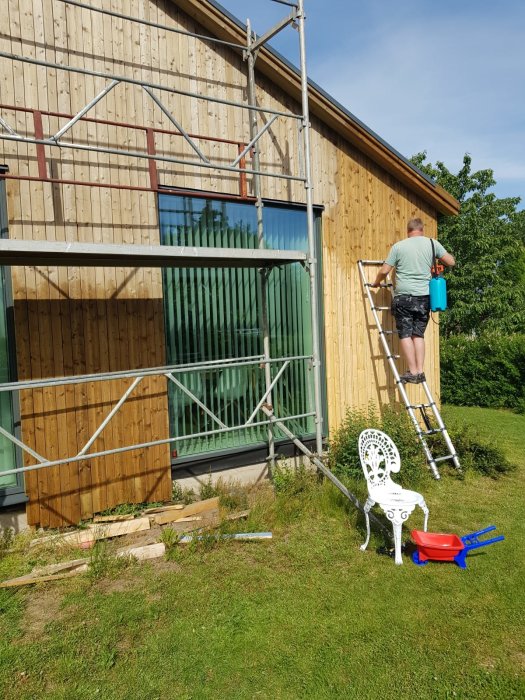 Person som använder en tryckspruta för att behandla träpanel på husgavel medan han står på en stege, omgivet av grönska och byggmaterial.