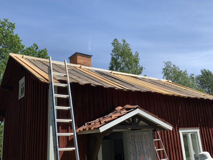 Delvis renoverat tak med nya läkter på en rödmålad stuga, stege mot taket, gammal takbeklädnad synlig på övrigt tak.