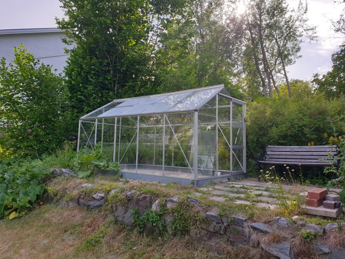 Ett litet begagnat växthus av glas och metall på en gräsmark omgivet av träd och buskar.