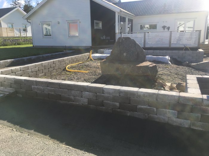 Delvis solbelyst altan med vitt räcke runt en 6x6 meter stor yta med murad kant, gräs och en sten i förgrunden.