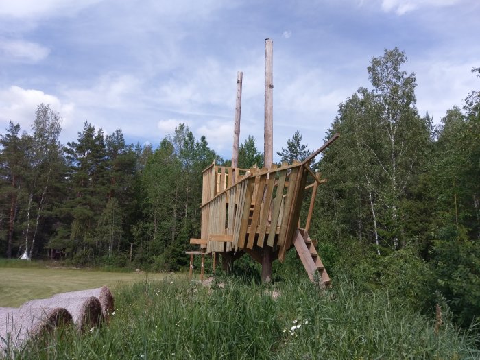 Träkonstruktion utomhus visar nybyggda trappor och ett delvis färdigt räcke med natur i bakgrunden.