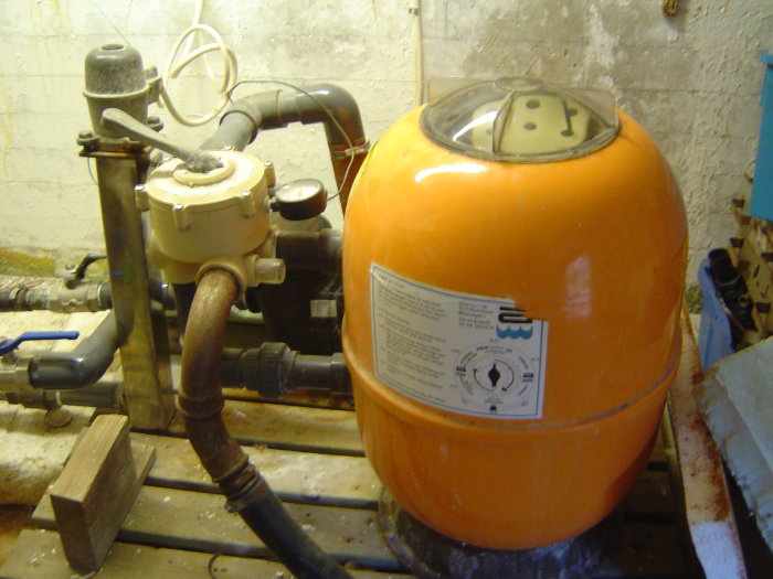 Gammal orange poolfiltertank med anslutna rör och pump i ett pumprum.