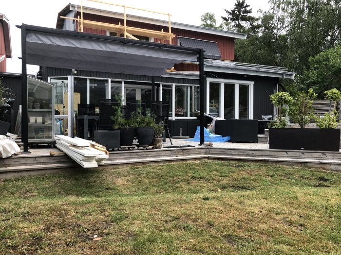 Nyinstallera pergola med tak fastskruvad i trädäck vid ett hus, med utomhusmöbler och växter runtomkring.