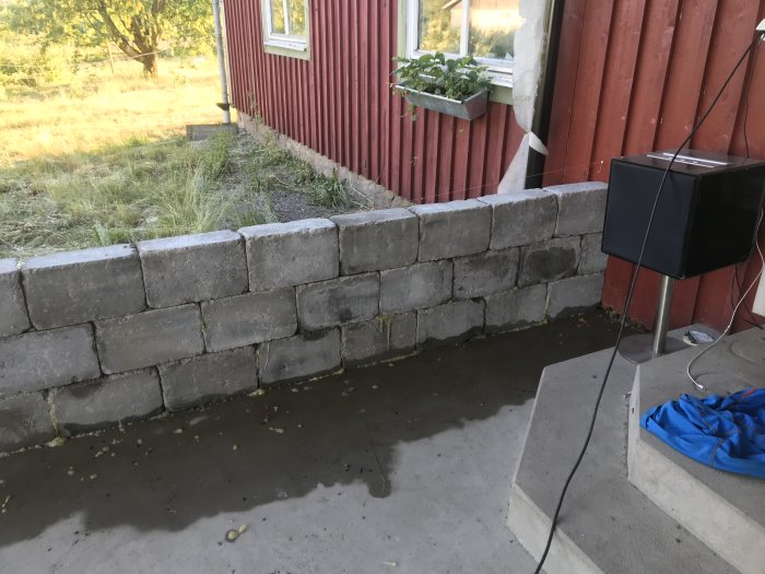 Nybyggd grå betongmur utomhus vid röd träbyggnad, med synlig elkabel och blå presenning.