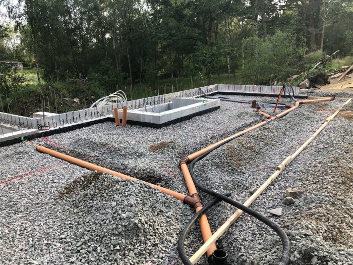 Grundarbeten för husbygge med synlig grund och rör på ett grusunderlag, omgivet av grönska.