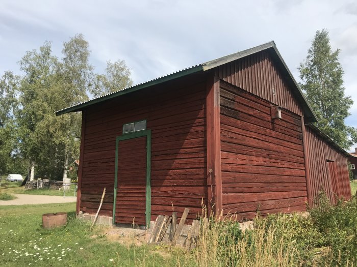 Gammal röd träbyggnad som tidigare användes som stall, omgiven av grönska.